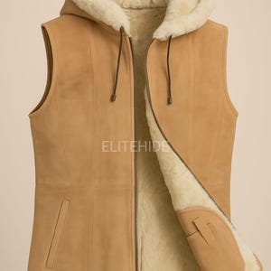 May include: A tan sleeveless vest with a hood, featuring a soft cream-colored shearling lining and a front zipper closure. The exterior is a smooth tan suede material with vertical seam detailing and two side pockets. The text ELITEHIDE is visible.