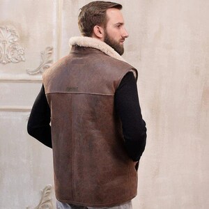 K&ouml;nnte beinhalten: Eine braune Lederweste mit einem Lammfellkragen. Die Weste hat einen Rei&szlig;verschluss und ist &auml;rmellos. Das Model tr&auml;gt die Weste &uuml;ber einem schwarzen Hemd und einer grauen Jeans.