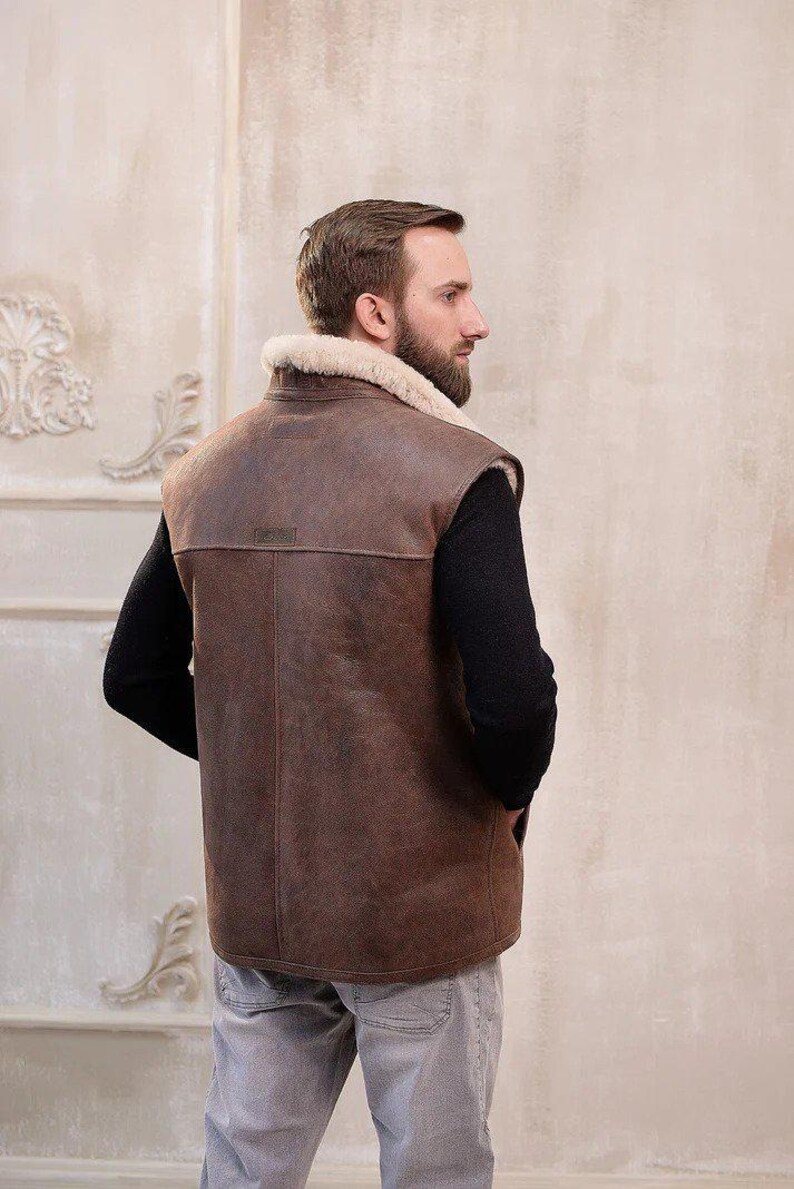 K&ouml;nnte beinhalten: Eine braune Lederweste mit einem Lammfellkragen. Die Weste hat einen Rei&szlig;verschluss und ist &auml;rmellos. Das Model tr&auml;gt die Weste &uuml;ber einem schwarzen Hemd und einer grauen Jeans.