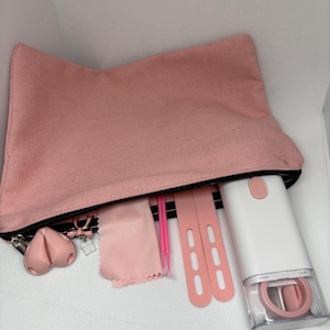 Peut inclure: Une trousse en toile rose avec une fermeture éclair noire, contenant divers outils de beauté. Comprend un charm en forme de cœur rose, un chiffon en microfibre rose, des applicateurs roses et un appareil blanc et rose.