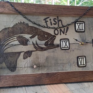 May include: A wooden clock with a fish design and the words "Fish On!" The clock has Roman numeral markings and a chain hanging from the top.