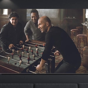 Könnte beinhalten: Drei Männer spielen Tischfußball in einer schwach beleuchteten Bar. Die Männer tragen Freizeitkleidung und konzentrieren sich auf das Spiel. Die Bar ist mit Holzmöbeln eingerichtet und hat ein Vintage-Feeling.