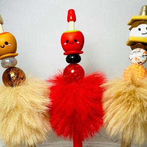 May include: Three decorative pen toppers with playful designs. One features a caramel apple, another a red apple, and the third a s'more. Each has a fluffy base and colourful beads. The toppers are approximately 15 cm tall.