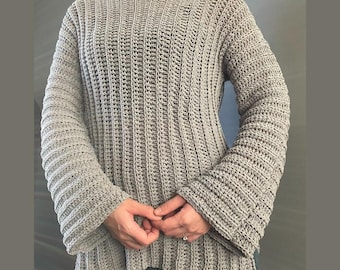 May include: A light grey, handmade crochet sweater with a ribbed texture. The sweater has a crew neck and long sleeves with flared cuffs. The garment is made of a soft, textured yarn.