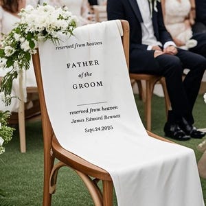 Puede incluir: Una silla de madera con tela blanca que dice "reserved from heaven, FATHER of the GROOM, James Edward Bennett, Sept.24.2025." La silla está decorada con flores blancas, en el contexto de una ceremonia de boda.