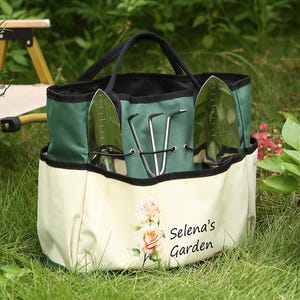 Peut inclure: Un sac fourre-tout de jardin vert et crème avec des poignées et des poches noires. Le sac contient des outils de jardinage et présente le texte "Selena's Garden" avec un motif floral. Le sac est sur l'herbe verte.