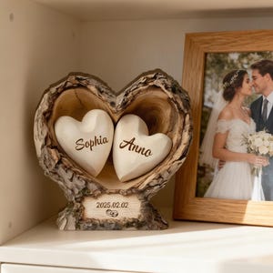 May include: A wooden heart-shaped ornament with two carved hearts, each inscribed with a name. The base is engraved with a date. A framed wedding photo is in the background. The ornament is on a white shelf.