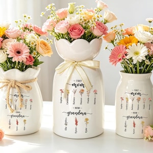 May include: Three white ceramic vases with floral arrangements. The vases are inscribed with "First Mom - Now Grandma" and names. The flowers are pink, yellow, and orange. One vase has a jute tie, another has a ribbon.