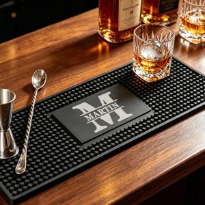 May include: A rectangular black rubber bar mat with a textured nub surface, featuring a central rectangular panel engraved with a large letter M and the name MARTIN. A stainless steel jigger and a long twisted bar spoon rest on the mat.