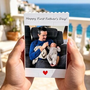 Puede incluir: Un bloque blanco con el texto "Happy First Father's Day!" y un coraz&oacute;n rojo. El bloque muestra una foto de un hombre y un beb&eacute;. El hombre lleva una camisa azul y sostiene al beb&eacute;. El beb&eacute; lleva un mono estampado.