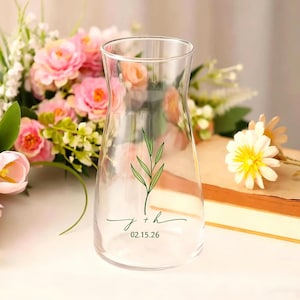 May include: Clear glass vase with a flared shape, featuring a green leaf design and the initials "j + h" with the date "02.15.26" in a delicate script. The vase is surrounded by pink flowers and a book.