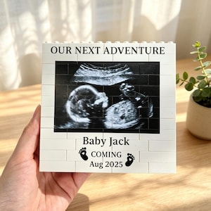 May include: A white brick-style block with a sonogram image, the text "OUR NEXT ADVENTURE", and the text "Baby Jack COMING Aug 2025" with footprints. The block is held in a hand.