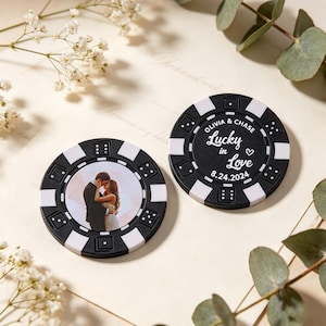 May include: Two black and white poker chips with a wedding theme. One chip features a photo of a couple, the other says "Olivia & Chase Lucky in Love 8.24.2024". The chips have a classic casino design.