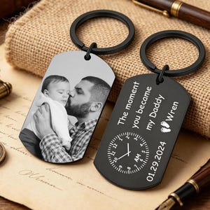 May include: Two black metal dog tag keychains with circular rings. One tag features a black and white photo of a person holding a baby. The other tag is engraved with the text The moment you became my Daddy, the name Wren, two footprints, a clock face set to 7 AM, and the date 01.29.2024.