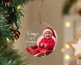 May include: A clear, round Christmas ornament with a photo of a baby in a Santa suit. The ornament has the text "Remy's 1st Christmas 2024" in elegant script. The ornament hangs from a gold string and is displayed on a Christmas tree.