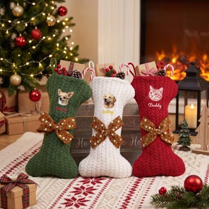 Puede incluir: Tres calcetines navideños con forma de hueso en tejido de punto trenzado verde oscuro, blanco y rojo. Cada calcetín presenta un retrato de mascota, un nombre bordado en cursiva y un lazo marrón con lunares. Están rellenos de cajitas de regalo.