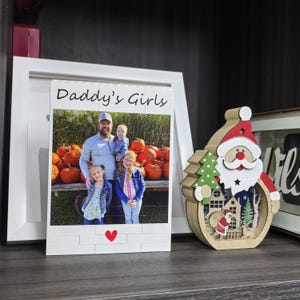 Puede incluir: Un marco de fotos de madera blanca con el texto "Daddy's Girls" y una foto de un hombre y tres ni&ntilde;as frente a calabazas. Un coraz&oacute;n rojo en la parte inferior. Decoraci&oacute;n de madera de Pap&aacute; Noel a la derecha.
