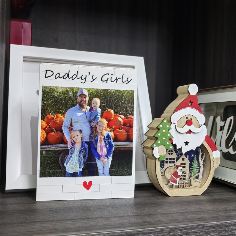 Puede incluir: Un marco de fotos de madera blanca con el texto "Daddy's Girls" y una foto de un hombre y tres ni&ntilde;as frente a calabazas. Un coraz&oacute;n rojo en la parte inferior. Decoraci&oacute;n de madera de Pap&aacute; Noel a la derecha.
