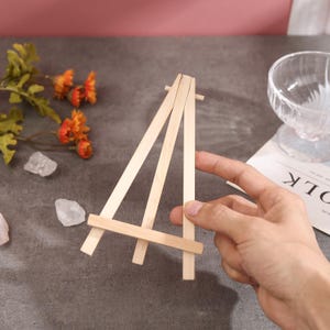 Puede incluir: Un peque&ntilde;o caballete de madera sin terminar, sostenido por una mano. El caballete est&aacute; hecho de madera de color claro y tiene un dise&ntilde;o simple en forma de A. Est&aacute; dise&ntilde;ado para sostener un lienzo peque&ntilde;o o una obra de arte. El fondo incluye flores y un cuenco de cristal.