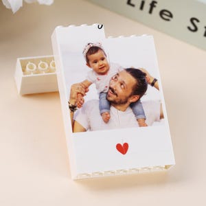 Puede incluir: Ladrillo Lego blanco con una fotograf&iacute;a impresa de una persona que lleva a un beb&eacute; sobre sus hombros. Un coraz&oacute;n rojo en la parte inferior. Otro ladrillo Lego blanco en el fondo.