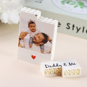 Puede incluir: Bloque de fotos blanco estilo Lego con una foto de padre e hijo. El bloque tiene un coraz&oacute;n rojo y las palabras "Daddy & Me" en una pieza separada. Un regalo personalizado.