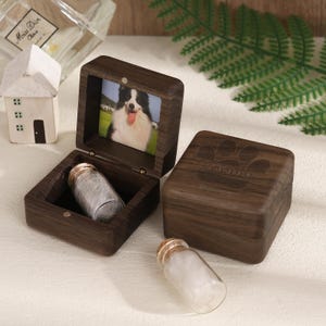 May include: A dark wood pet urn with a photo of a dog inside. The urn is open, revealing a small glass vial filled with white material. A second closed urn with a paw print design and the name "Cooper" is also present.