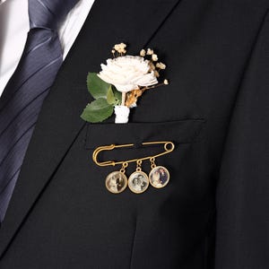 Puede incluir: Un broche de imperdible dorado con tres dijes circulares, cada uno con una pequeña fotografía en blanco y negro de una pareja. El broche está sujeto al bolsillo de una chaqueta de traje negra, que también lleva un boutonniere floral blanco.