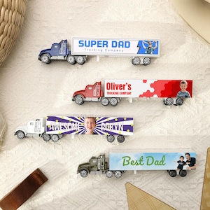 May include: Four toy semi-trucks with personalized trailer designs. The trucks are blue, red, white, and green. The trailers feature the phrases "SUPER DAD", "Oliver's", "AWESOME ROBYN", and "Best Dad", along with images.