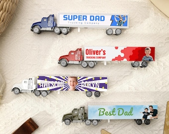 May include: Four toy semi-trucks with personalized trailer designs. The trucks are blue, red, white, and green. The trailers feature the phrases "SUPER DAD", "Oliver's", "AWESOME ROBYN", and "Best Dad", along with images.