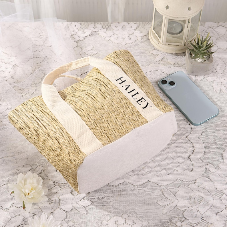 Peut inclure: Un sac fourre-tout beige tiss&eacute; avec des accents et des poign&eacute;es blancs. Le mot "HAILEY" est imprim&eacute; en noir sur la bande blanche. Un smartphone bleu clair est &agrave; proximit&eacute;. Le sac est sur une surface en dentelle blanche.