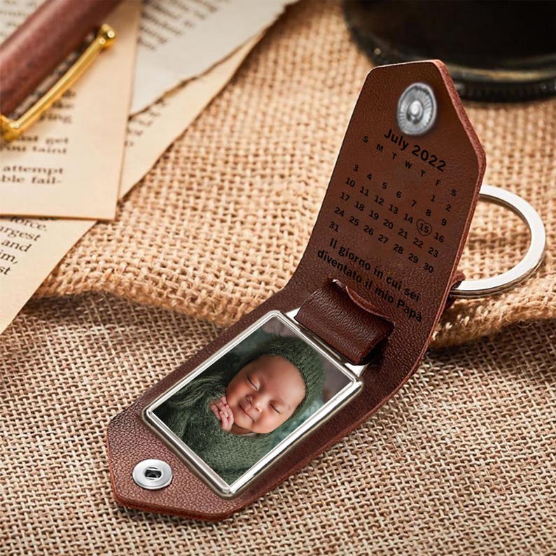 May include: A brown leather keychain with a photo insert and a calendar engraved with the text "July 2022". The calendar has the number 15 circled. The text "Il giorno in cui sei diventato il mio Papa" is also engraved on the keychain.