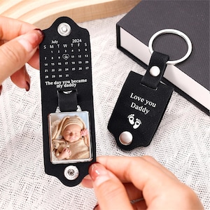 May include: Black leather keychain with a silver photo frame and a calendar engraved with the text "The day you became my daddy". The photo frame contains a picture of a baby. The keychain also has a black leather tag with the text "Love you Daddy" and a small footprint design.