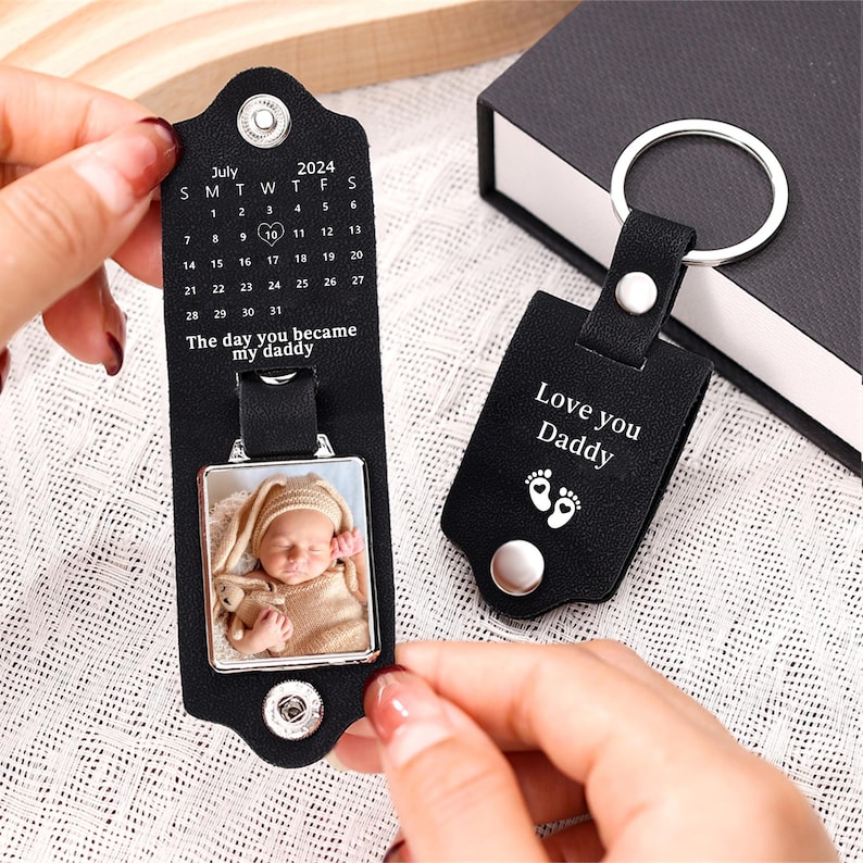 May include: Black leather keychain with a silver photo frame and a calendar engraved with the text "The day you became my daddy". The photo frame contains a picture of a baby. The keychain also has a black leather tag with the text "Love you Daddy" and a small footprint design.