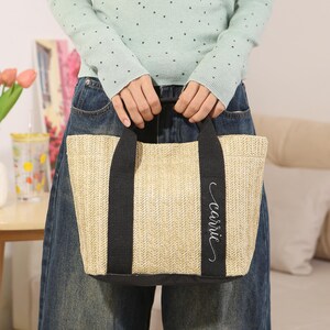 Peut inclure: Un sac fourre-tout beige tiss&eacute; avec des poign&eacute;es et une base noires. Le sac porte le nom "carrie" en &eacute;criture cursive sur le c&ocirc;t&eacute;. Le sac est tenu par une personne portant un pull vert clair et un jean bleu.