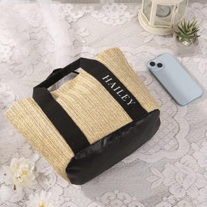 Peut inclure: Un sac fourre-tout en paille tress&eacute;e avec des accents noirs et le nom "HAILEY" imprim&eacute; en blanc. Le sac a une base et des poign&eacute;es noires. Un smartphone bleu clair et une petite plante succulente sont en arri&egrave;re-plan.