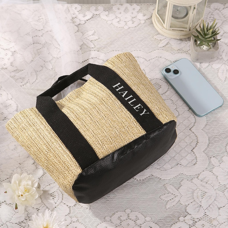 Peut inclure: Un sac fourre-tout en paille tress&eacute;e avec des accents noirs et le nom "HAILEY" imprim&eacute; en blanc. Le sac a une base et des poign&eacute;es noires. Un smartphone bleu clair et une petite plante succulente sont en arri&egrave;re-plan.