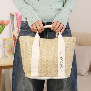 Peut inclure: Un sac fourre-tout en paille tress&eacute;e avec des poign&eacute;es et des accents en toile blanche. Le sac porte le nom "Carrie" en &eacute;criture cursive sur le devant. Le sac est tenu par une personne portant un pull bleu clair et un jean bleu.