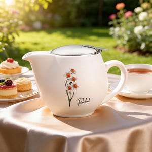 Könnte beinhalten: Cremefarbene Keramik-Teekanne mit Edelstahl-Deckel und Griff. Die Teekanne hat ein Blumendesign mit orangefarbenen und weißen Blumen und dem Namen "Rachel". Eine Teetasse und Gebäck stehen ebenfalls auf dem Tisch.