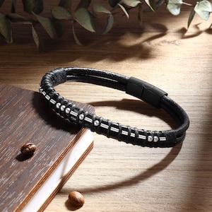 May include: Black leather bracelet with silver-coloured detailing and a magnetic clasp. The bracelet has a braided design with small, round silver beads. It is displayed on a wooden surface.