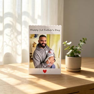 Puede incluir: Un marco de fotos de bloque blanco con el texto "Happy 1st Father's Day" y un coraz&oacute;n rojo. El marco contiene una foto de un hombre sosteniendo a un beb&eacute;. Una peque&ntilde;a planta en maceta est&aacute; a la derecha.