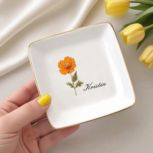 Può includere: Un vassoio quadrato in ceramica bianca con bordo dorato. Il piatto presenta un'illustrazione di un fiore arancione e il nome "Kristin" in corsivo nero. Il piatto è tenuto da una persona con lo smalto giallo.