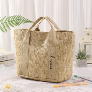Peut inclure: Un sac fourre-tout beige tiss&eacute; avec des poign&eacute;es robustes et le nom "hanna" en &eacute;criture cursive sur le c&ocirc;t&eacute;. Le sac a une forme rectangulaire et est fait d'un mat&eacute;riau d'aspect naturel.