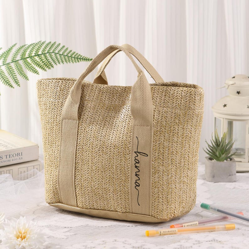 Peut inclure: Un sac fourre-tout beige tiss&eacute; avec des poign&eacute;es robustes et le nom "hanna" en &eacute;criture cursive sur le c&ocirc;t&eacute;. Le sac a une forme rectangulaire et est fait d'un mat&eacute;riau d'aspect naturel.