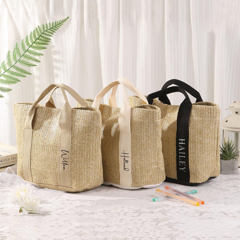 Peut inclure: Trois sacs fourre-tout en paille tress&eacute;e avec des anses en tissu. Un sac a une anse noire et le nom "Hailey" imprim&eacute; sur une bande verticale. Les deux autres sacs ont des anses beiges.