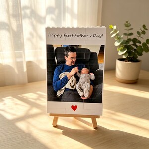 Puede incluir: Un marco de fotos blanco estilo bloque con el texto "Happy First Father's Day!" y un coraz&oacute;n rojo. El marco muestra una foto de una persona sosteniendo a un beb&eacute;. El marco se muestra en un peque&ntilde;o caballete de madera.