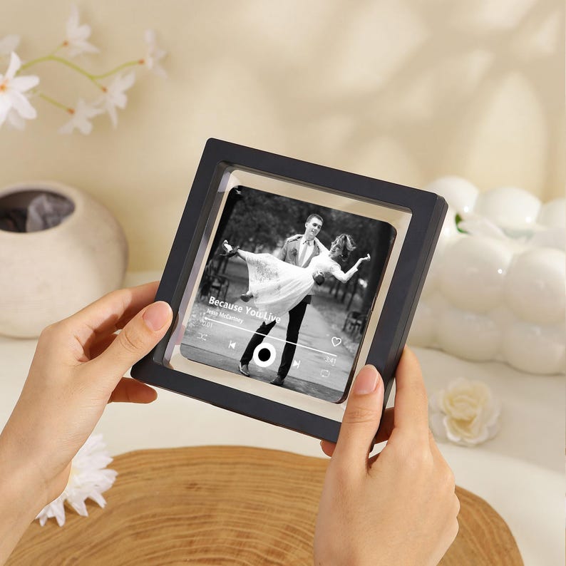May include: A black and white photo of a couple dancing, displayed in a black square frame. The frame is held by hands. The photo has the text "Because You Live" and a music player interface.