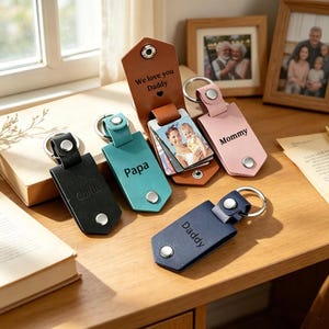 May include: A selection of leather keyrings in black, teal, pink, and navy. Each keyring has a silver metal ring and a snap closure. Some keyrings are inscribed with "Carlos", "Papa", "Mommy", and "Daddy". One keyring has the text "We love you Daddy".