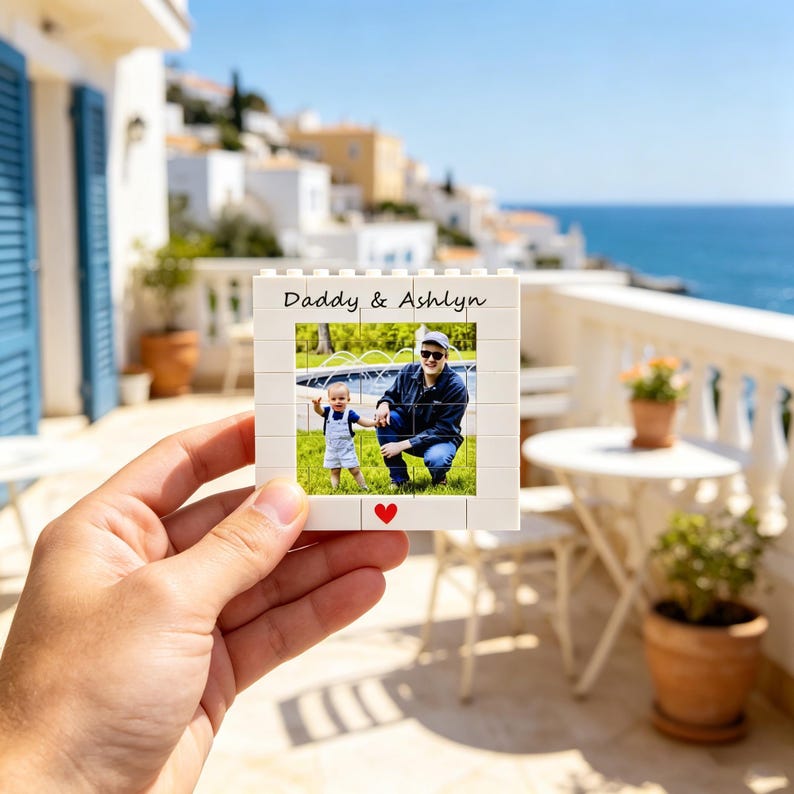 Puede incluir: Un marco de fotos blanco con una foto de un padre y un ni&ntilde;o, con el texto "Daddy & Ashlyn" en la parte superior y un coraz&oacute;n rojo en la parte inferior. El marco se sostiene en una mano contra un fondo esc&eacute;nico al aire libre.
