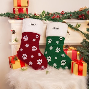 May include: Two personalized Christmas stockings with white paw print designs. The stocking on the left is red with the name "Elise" embroidered on the cuff. The stocking on the right is green with the name "Elise" embroidered on the cuff.