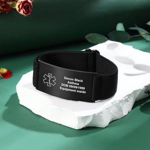 May include: Black medical alert bracelet with a black plate. The plate has a medical symbol and the following text: Devon Black, Asthma, DOB 09/09/1999, Equipment inside. The bracelet is on a white surface.